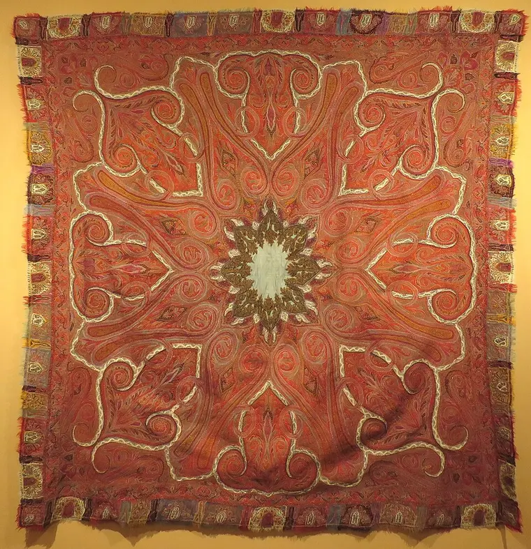 A square red Kashmir shawl in a museum, with a pale star-shaped centre, swirling paisleys and leaves, and a patchwork border.