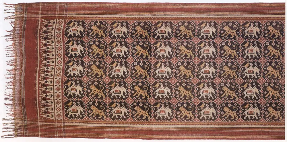 A long silk cloth with fringed ends, patterned in a grid of white elephants with riders and golden lions, all with slightly blurred edges.