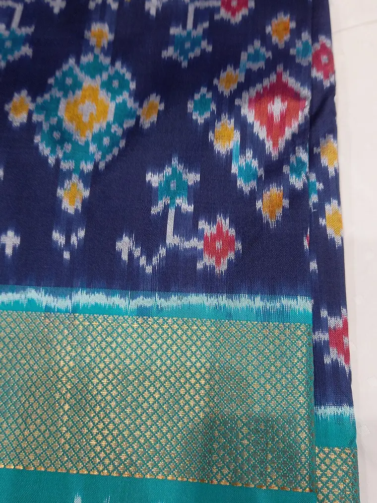 A navy silk sari with diamond and flower shapes in red, yellow and teal, their edges blurred, above a teal border with a gold zari grid.