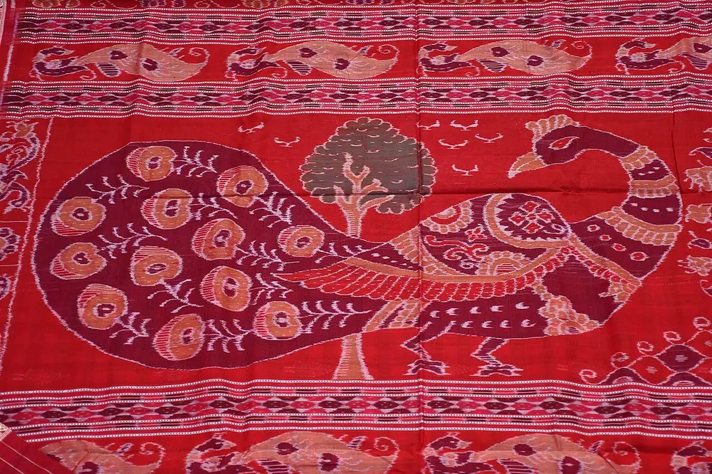The pallu of a red Sambalpuri sari, with a large peacock and a tree in maroon and beige, their outlines softly blurred, between bands of small motifs.